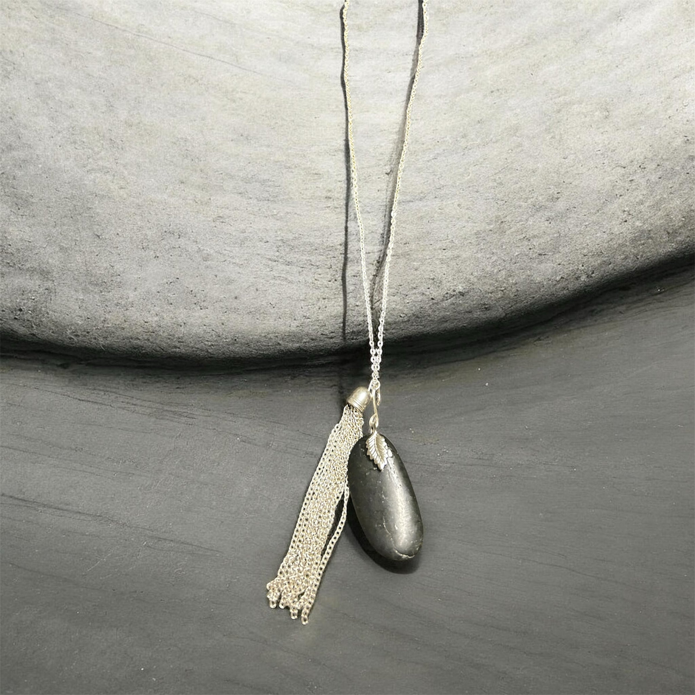 M.C’s decorative pendant necklace featuring a smooth dark river stone and a shimmering silver-tone tassel on a thin chain.